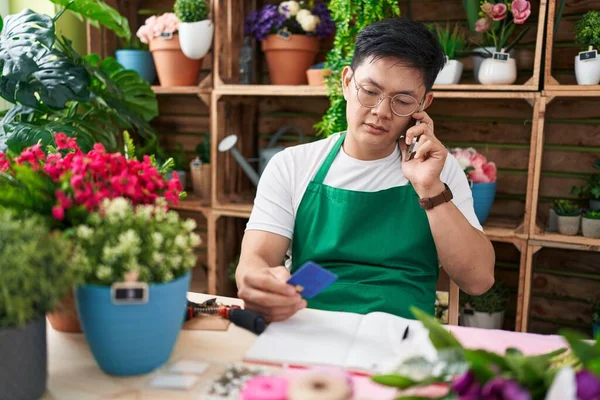 Young chinese man florist talking on smartphone holding credit card at flower shop