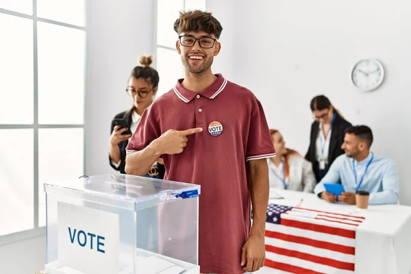 Young american voter man smiling happy pointing with finger to usa badge at vote center.