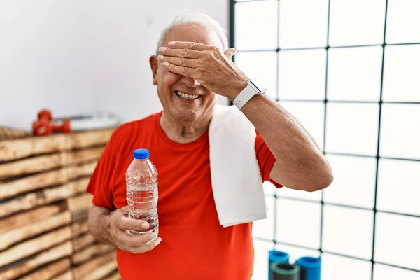 Senior man wearing sportswear and towel at the gym smiling and laughing with hand on face covering eyes for surprise. blind concept.