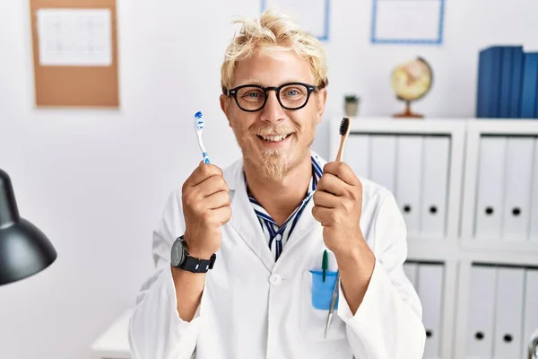 Young Blond Dentist Man Working Dentist Clinic Holding Toothbrushes Smiling — Foto Stock