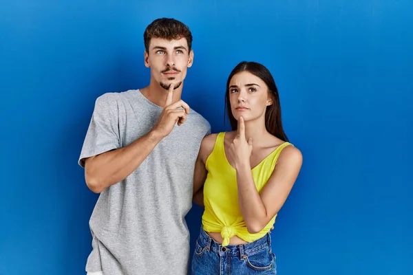 Young hispanic couple standing together over blue background thinking concentrated about doubt with finger on chin and looking up wondering