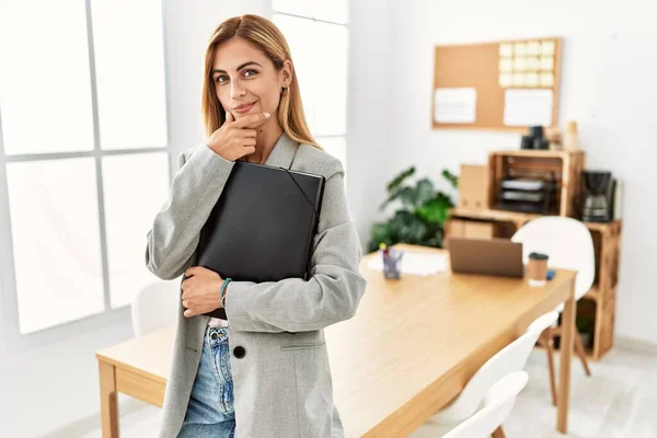 Blonde business woman at the office looking confident at the camera smiling with crossed arms and hand raised on chin. thinking positive.