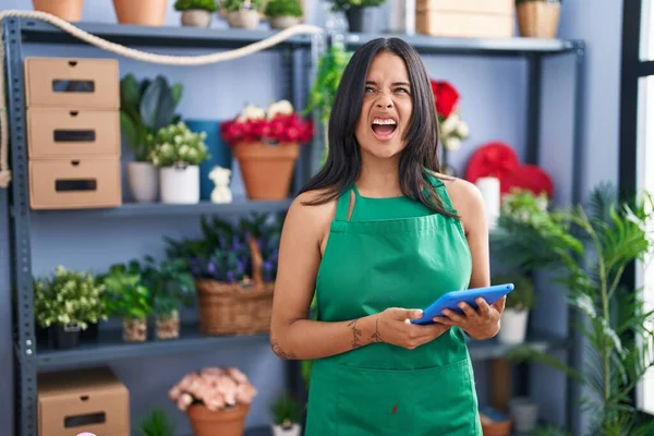Brunette woman working at florist shop holding tablet angry and mad screaming frustrated and furious, shouting with anger looking up.