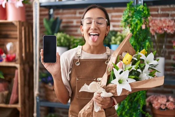 若いヒスパニック系の女性が花屋で働いていますスマートフォンの画面を示す面白い表現で幸せ舌を突き出す — ストック写真