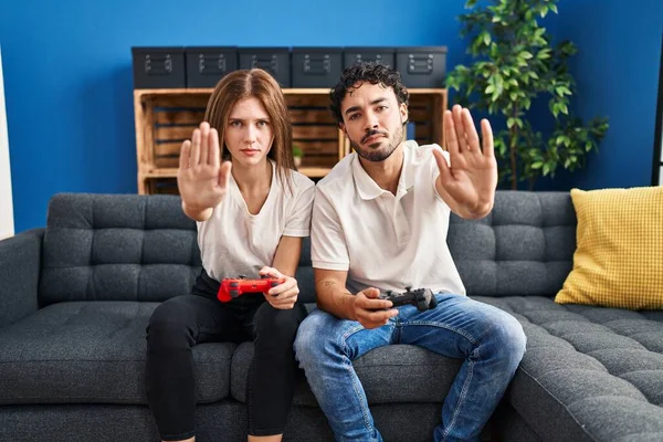 Young couple playing video game holding controller at home with open hand doing stop sign with serious and confident expression, defense gesture
