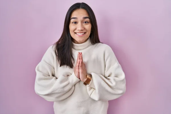 Young south asian woman standing over pink background praying with hands together asking for forgiveness smiling confident.