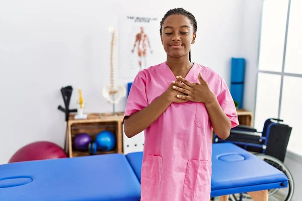 Young african american woman working at pain recovery clinic smiling with hands on chest with closed eyes and grateful gesture on face. health concept.