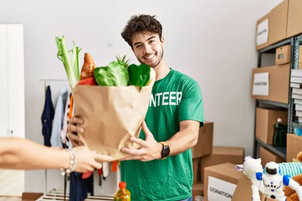 Young Hispanic Man Wearing Volunteer Uniform Giving Groceries Paper Bag — Stockfoto