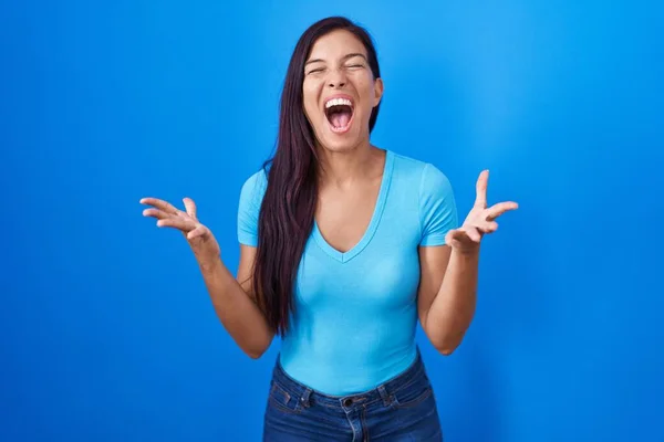 腕を上げて 興奮して叫んで目を閉じて成功のために狂気と狂気を祝う青い背景の上に立って若いヒスパニック系の女性 勝者のコンセプト — ストック写真