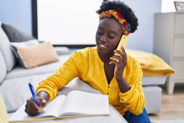 Young african american woman writing on notebook talking on the smartphone at home