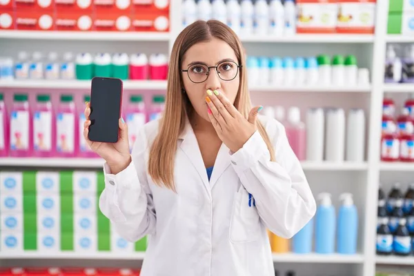 年轻的金发女子在药店工作 手拿着智能手机屏幕捂住嘴 很震惊 很害怕犯错 惊讶的表情 — 图库照片