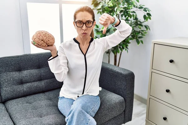 Young hispanic woman holding brain working at psychology clinic with angry face, negative sign showing dislike with thumbs down, rejection concept