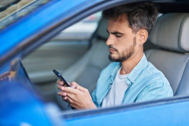 Young hispanic man using smartphone sitting on car at street