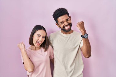 Young hispanic couple together over pink background very happy and excited doing winner gesture with arms raised, smiling and screaming for success. celebration concept. 
