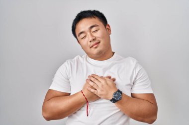 Young chinese man standing over white background smiling with hands on chest with closed eyes and grateful gesture on face. health concept. 