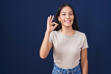 Mavi arka planda duran genç İspanyol kadın gülümsüyor ve el ve parmaklarıyla 