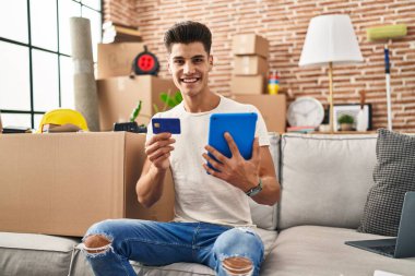 Young hispanic man using touchpad and credit card sitting on sofa at new home