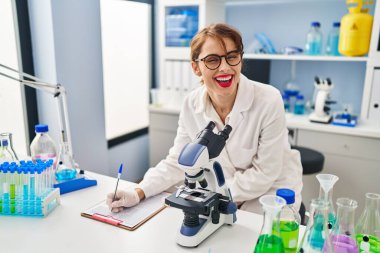 Genç beyaz kadın bilim adamı üniforması giyiyor. Laboratuvarda panoya ve mikroskoba yazı yazıyor.