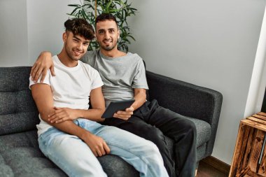 Two hispanic men couple using touchpad hugging each other at home