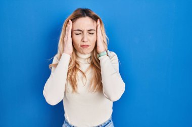Young caucasian woman standing over blue background with hand on head, headache because stress. suffering migraine. 