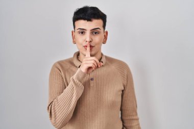 Non binary person standing over isolated background asking to be quiet with finger on lips. silence and secret concept. 