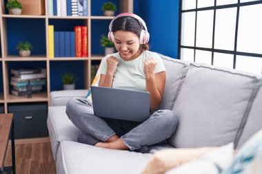 Hispanic young woman using laptop at home wearing headphones very happy and excited doing winner gesture with arms raised, smiling and screaming for success. celebration concept. 