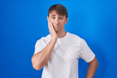 Caucasian blond man standing over blue background looking stressed and nervous with hands on mouth biting nails. anxiety problem. 