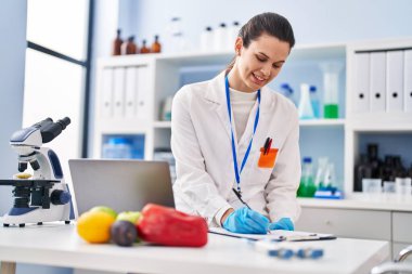 Young beautiful hispanic woman scientist using laptop writing on document at laboratory