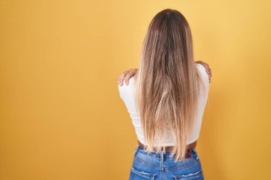 Young blonde woman standing over yellow background wearing headphones hugging oneself happy and positive from backwards. self love and self care 