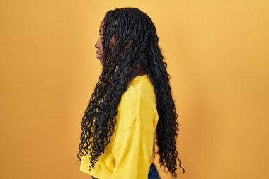 Sarı arka planda dikilen Afrikalı kadın, yan tarafa bakıyor, rahat poz veriyor kendine güvenen bir gülümsemeyle doğal bir yüz ifadesi sergiliyor.. 