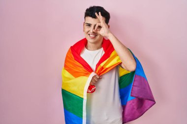 Non binary person holding rainbow lgbtq flag smiling happy doing ok sign with hand on eye looking through fingers 