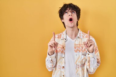 Yazlık tişört giyen genç adam şaşırmış ve şaşırmış bir şekilde yukarı bakıyor ve parmaklarıyla işaret ediyor ve kollarını kaldırıyor.. 