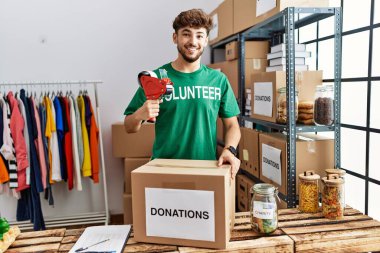 Young arab man wearing volunteer uniform packing donations box at charity center