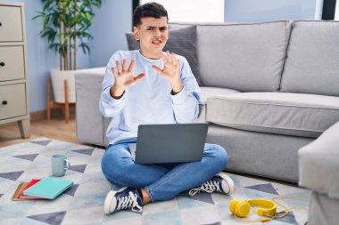 Non binary person studying using computer laptop sitting on the floor afraid and terrified with fear expression stop gesture with hands, shouting in shock. panic concept. 