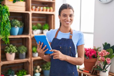 Brunette young woman working at florist shop holding tablet looking positive and happy standing and smiling with a confident smile showing teeth 