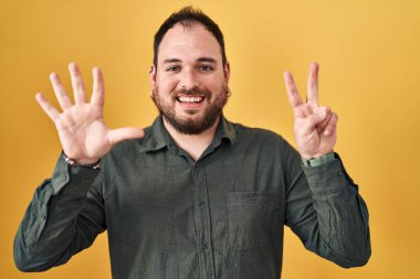 Plus size hispanic man with beard standing over yellow background showing and pointing up with fingers number seven while smiling confident and happy. 