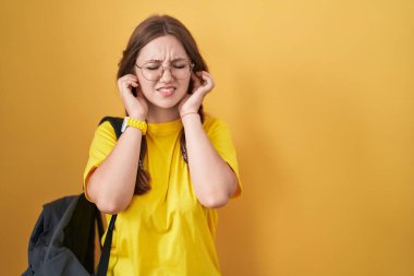 Sarı arka planda öğrenci çantası takan beyaz bir kadın yüksek sesli müzik sesi için kulaklarını parmaklarıyla kapatıyor. sağır kavramı. 