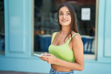 Genç, güzel, İspanyol bir kadın sokakta akıllı telefon kullanarak gülümsüyor.