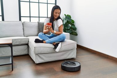 Genç Latin kadın evde akıllı robot süpürgesi kullanarak kahve içti.