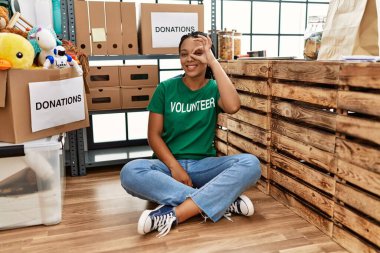 Young african american woman wearing volunteer t shirt at donations stand doing ok gesture with hand smiling, eye looking through fingers with happy face. 