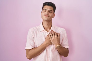 Pembe arka planda duran genç İspanyol adam elleri göğsünde, gözleri kapalı ve yüzünde minnet dolu bir jest ile gülümsüyor. sağlık kavramı. 