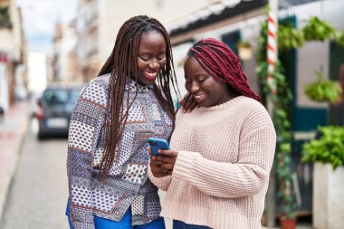 African american women friends smiling confident using smartphone at street