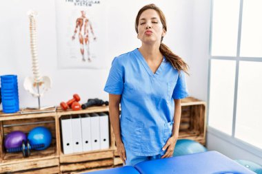 Middle age hispanic physiotherapist woman working at pain recovery clinic looking at the camera blowing a kiss on air being lovely and sexy. love expression. 