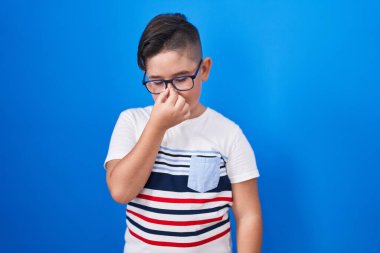 Mavi arka planda dikilen genç İspanyol çocuk yorgun burnu ve gözleri ovuşturup yorgunluk ve baş ağrısı hissediyor. Stres ve hüsran kavramı. 