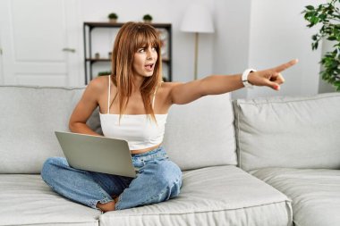 İspanyol kadın evdeki kanepede oturmuş dizüstü bilgisayarla işaret ediyor. Önünü işaret ediyor. Şaşırmış, ağzı açık, şaşırmış bir ifade. 