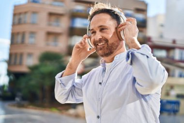 Genç, beyaz bir adam gülerek sokakta müzik dinliyor.