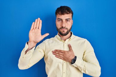 Handsome hispanic man standing over blue background swearing with hand on chest and open palm, making a loyalty promise oath 