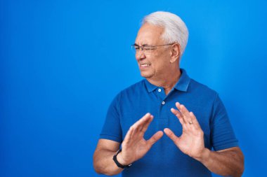 Mavi arka planda duran gri saçlı orta yaşlı bir adam tiksinti reaksiyonu yüzünden tiksinti ve korku dolu bir ifade takınıyor. Eller havada. 