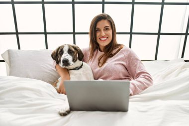 Yatak odasında dizüstü bilgisayar kullanan, köpeğini kucaklayan genç bir kadın.