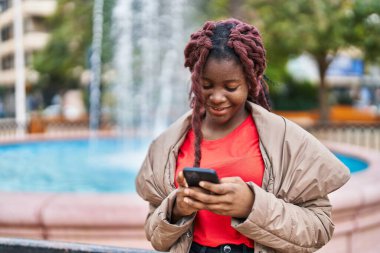 Afro-Amerikalı kadın parkta akıllı telefon kullanarak gülümsüyor.